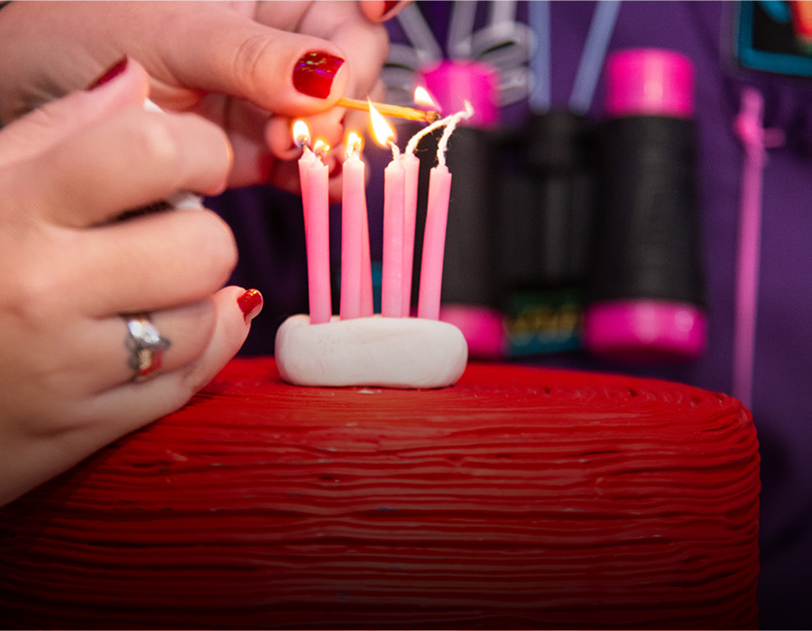 A close-up shot of two hands with red nail polish using a lit match to light several pink birthday candles arranged in a white candle holder on top of a red frosted cake.
