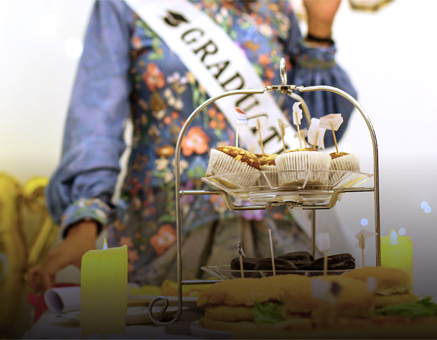 A graduate with a "Graduate" sash stands behind a tiered serving tray of food.