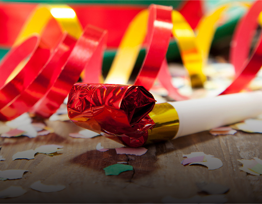 A close-up of a red and gold party noisemaker with red and yellow curled streamers extending from it. It rests on a surface covered in colorful confetti.